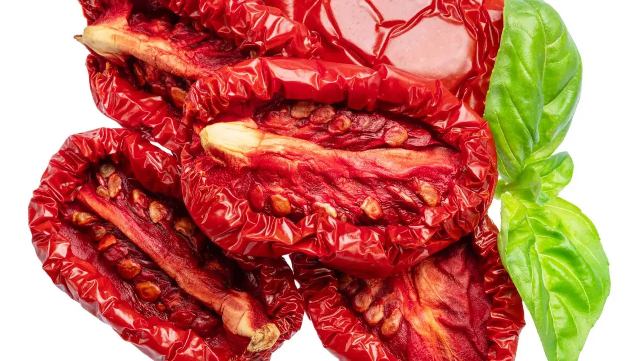 Tomates secos al horno, arrugados y rojos, con semillas visibles, junto a una hoja de albahaca verde fresca.