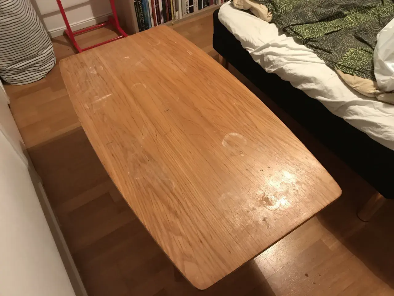 Ett slitet soffbord i trä med fläckar, redo att måla om. Bredvid syns en säng med vitt och grönt överkast.