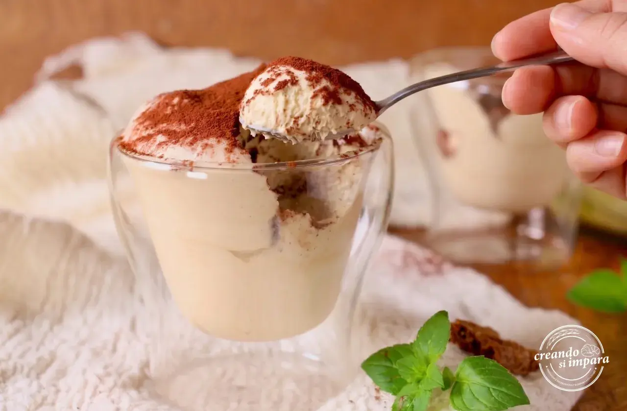 Un cucchiaino preleva una porzione generosa di soffice mousse al caffè, cosparsa di cacao, da un bicchiere di vetro.