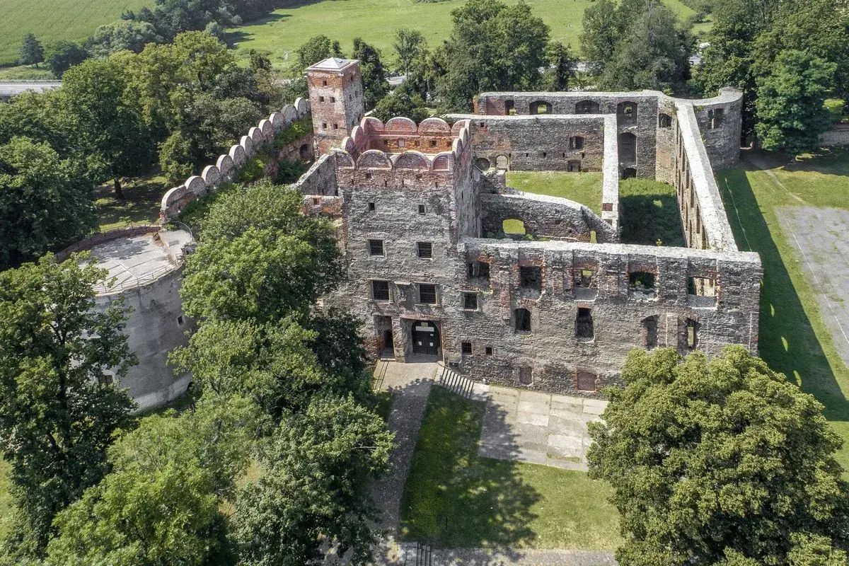 Ruiny zamku w Ząbkowicach Śląskich, z charakterystyczną okrągłą basteją i ceglanymi murami, otoczone zielenią.