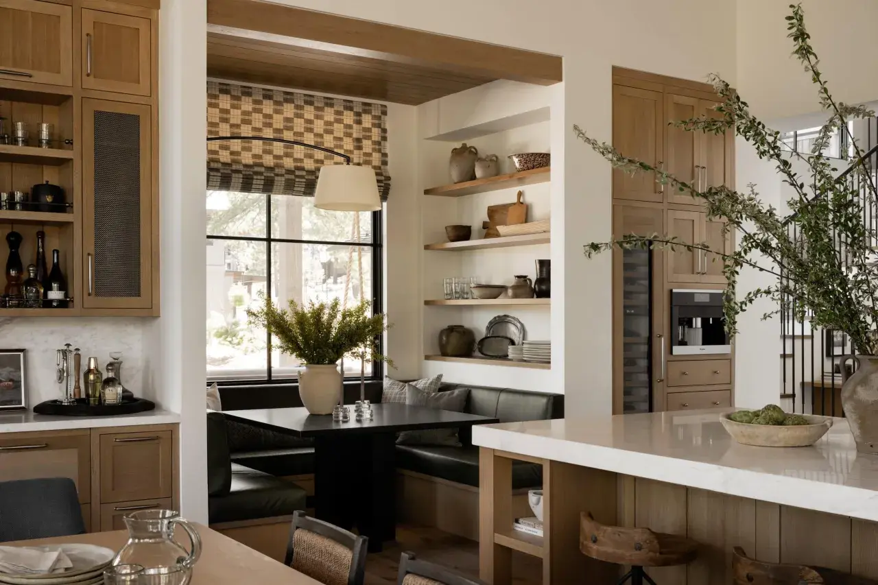 A cozy breakfast nook built in with a dark table, plush seating, and a large window. Shelves display rustic pottery and kitchenware.