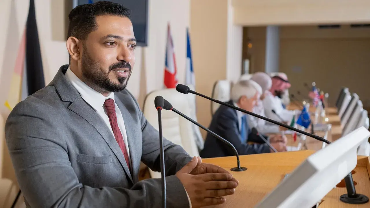 A man speaks at a podium, perhaps sharing insights from his pursuit of best online MPA programs, as others listen at a long table with flags.