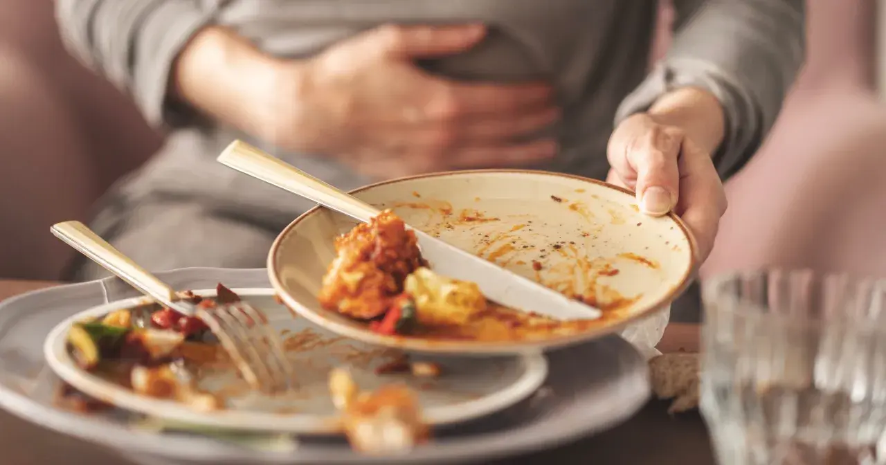 Osoba trzyma brudny talerz, czując mdłości po jedzeniu. Na stole resztki jedzenia i sztućce.