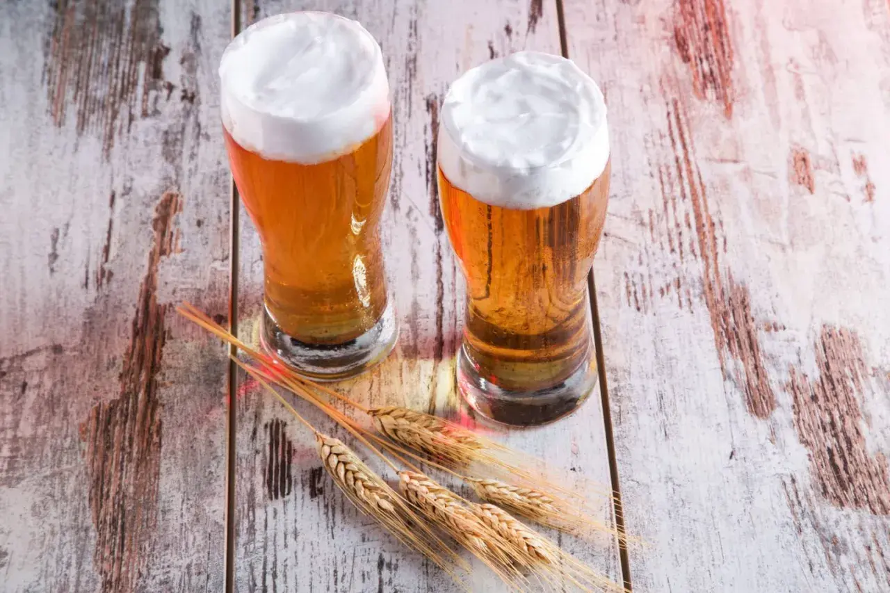 Dwa kufle złocistego piwa, przypominającego red ale, z obfitą pianką, stoją na drewnianym blacie obok kłosów zboża.