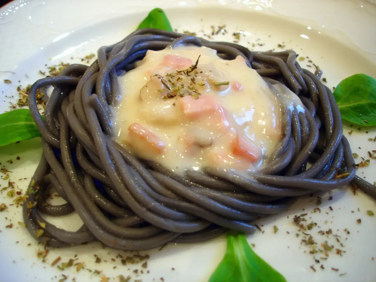 Espaguetis negro con nata, trocitos de jamón y hierbas aromáticas, servido en un plato blanco.