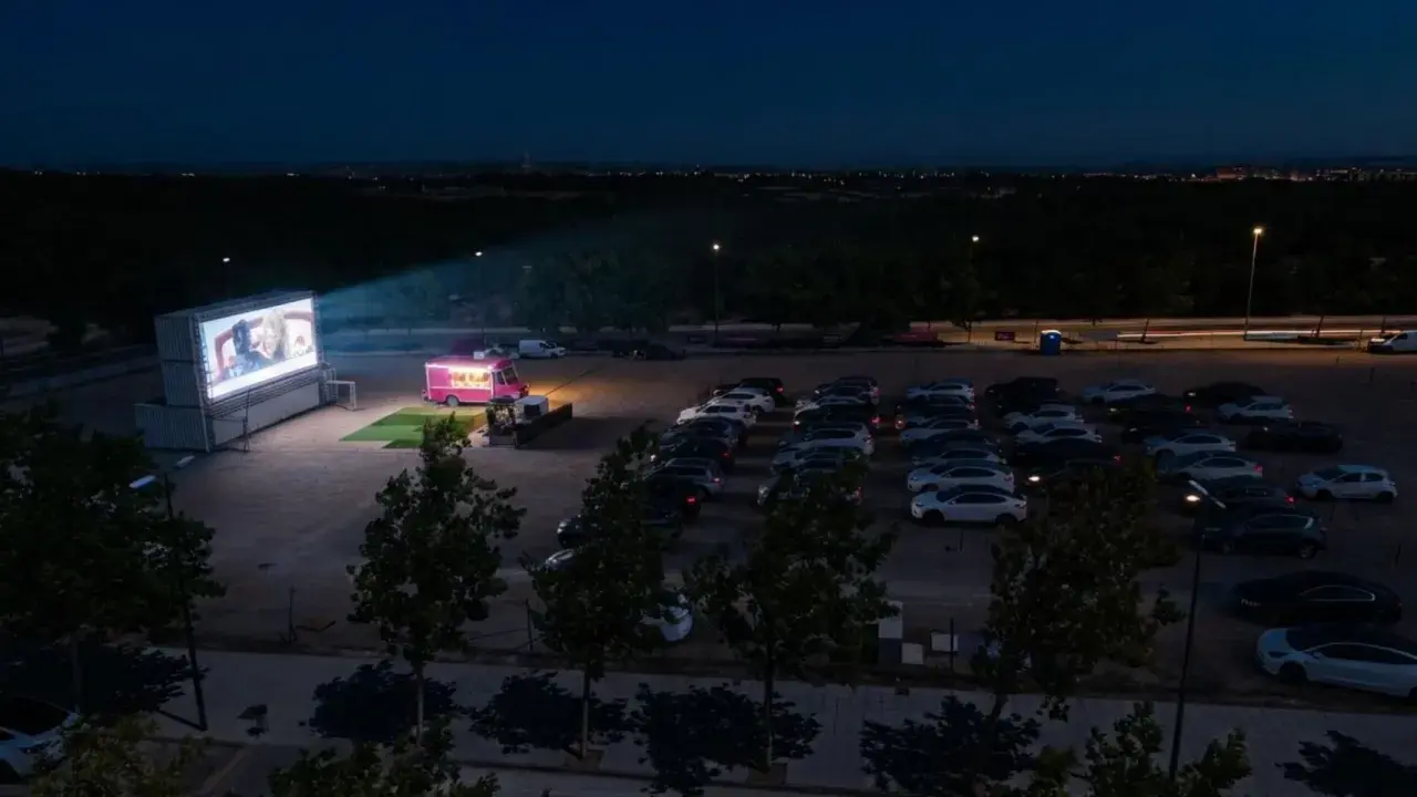 Noche de cine de verano Zaragoza. Coches aparcados frente a una gran pantalla donde proyectan una película, con un camión de comida rosa al lado.