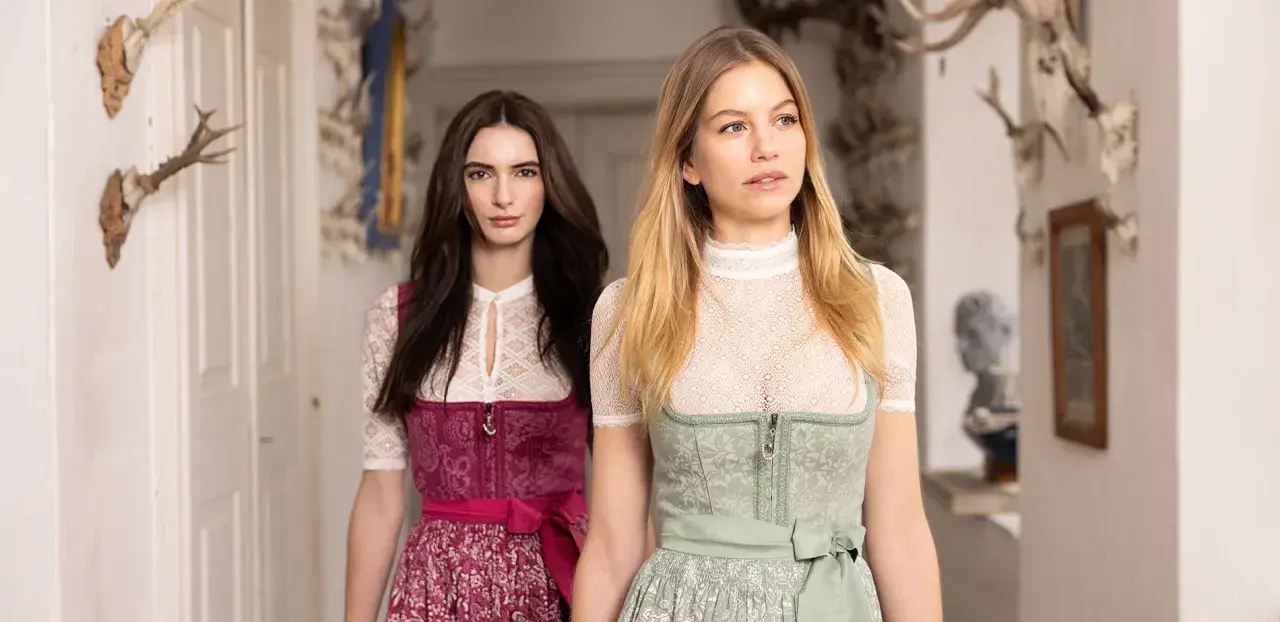Zwei Frauen in Tracht, eine mit dunkelbraunem, die andere mit blondem Haar, präsentieren elegante dirndlschürzen lang.