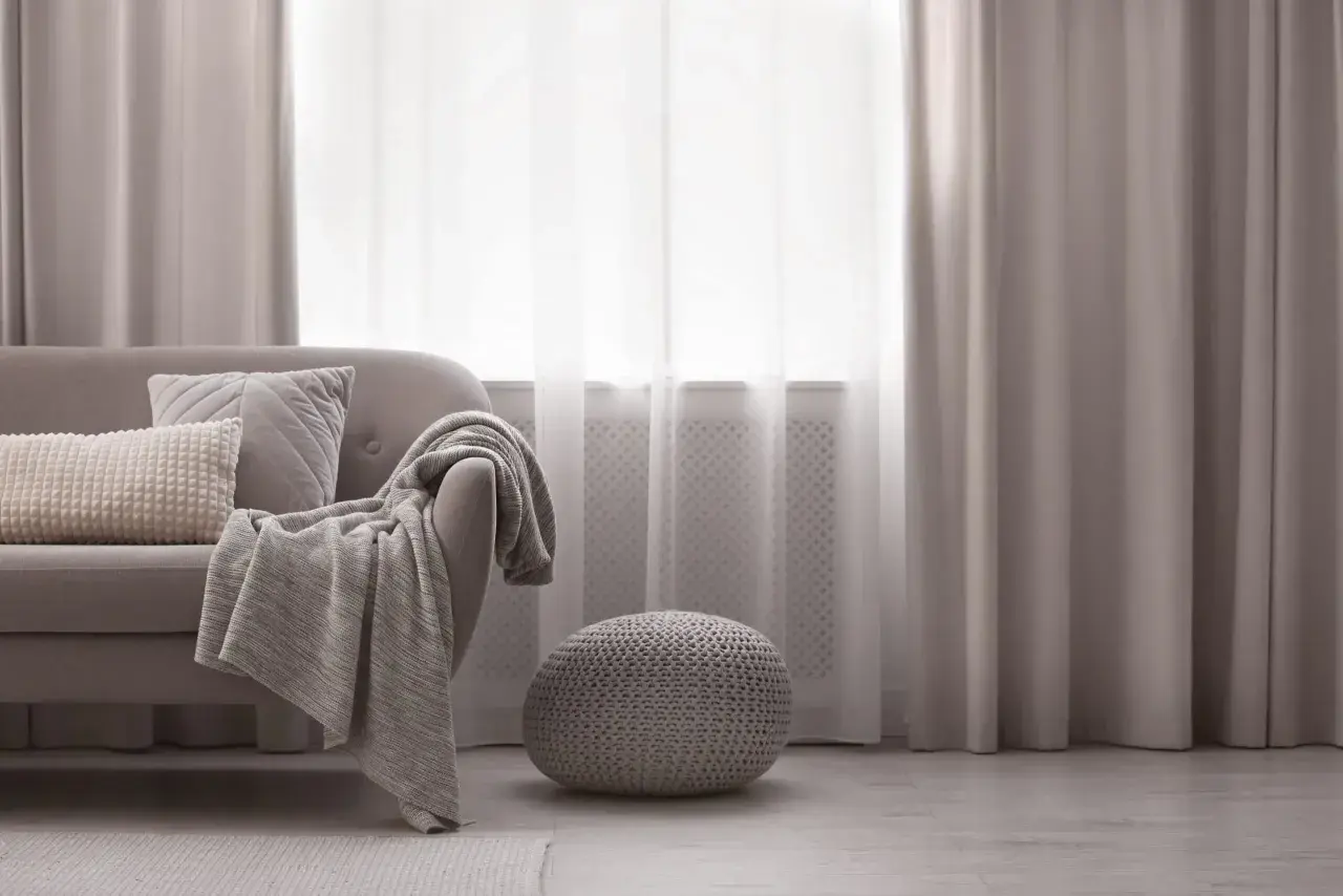 Miękka sofa z poduszkami i pledem, obok okrągła pufa. Elegancka dekoracja okien z zasłonami tworzy przytulną atmosferę.