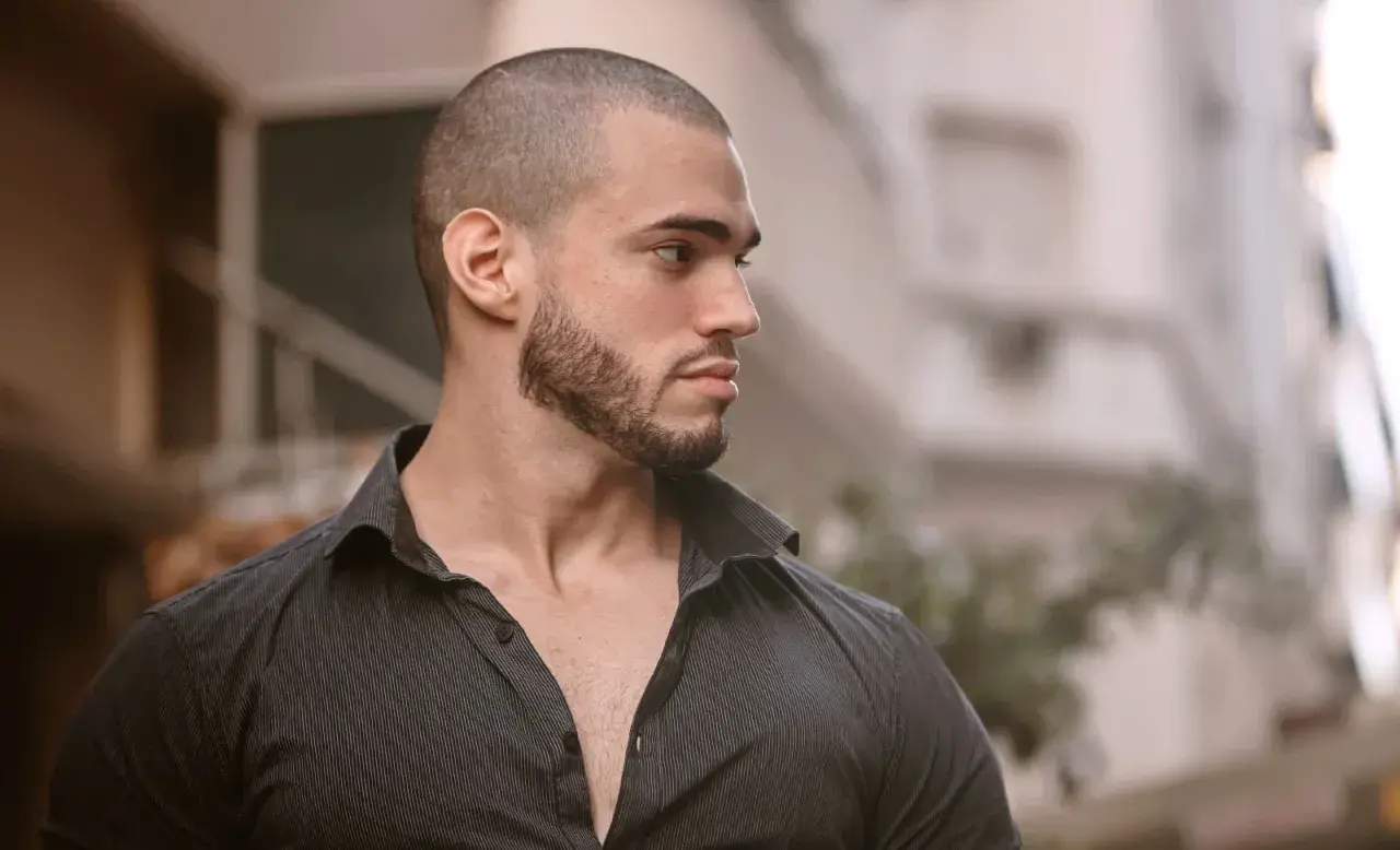 Hombre con buzz cut con entradas, barba cuidada y camisa oscura, mirando hacia un lado con expresión pensativa.