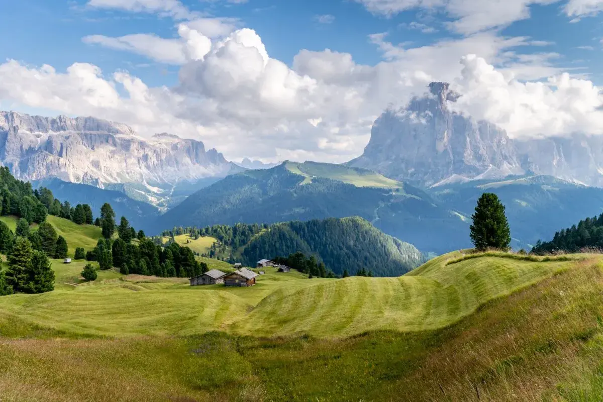 Zielone wzgórza i drewniane chatki w Val Gardena, z majestatycznymi Dolomitami w tle.