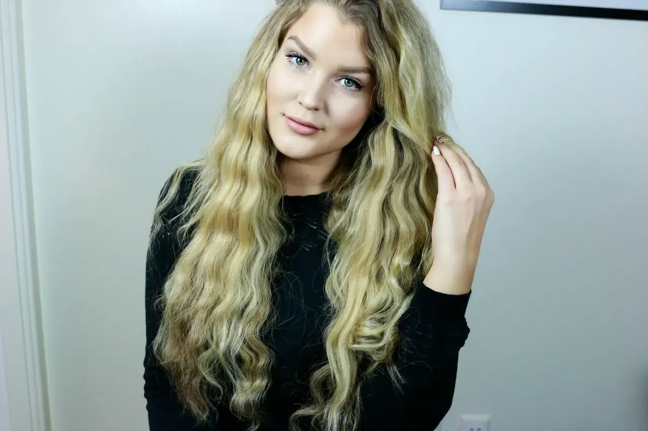 En ung kvinna med långt, blont, vågigt hår som rör vid det. Hon har gröna ögon och ljus hy. Hon bär en svart tröja. Frågan "hur får man lent hår" kan besvaras med denna look.
