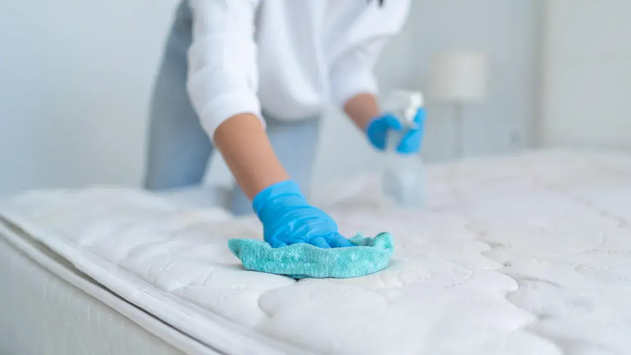 Persona con guantes azules limpia un colchón con un paño turquesa y spray. Aprende como limpiar colchones en casa para un descanso higiénico.
