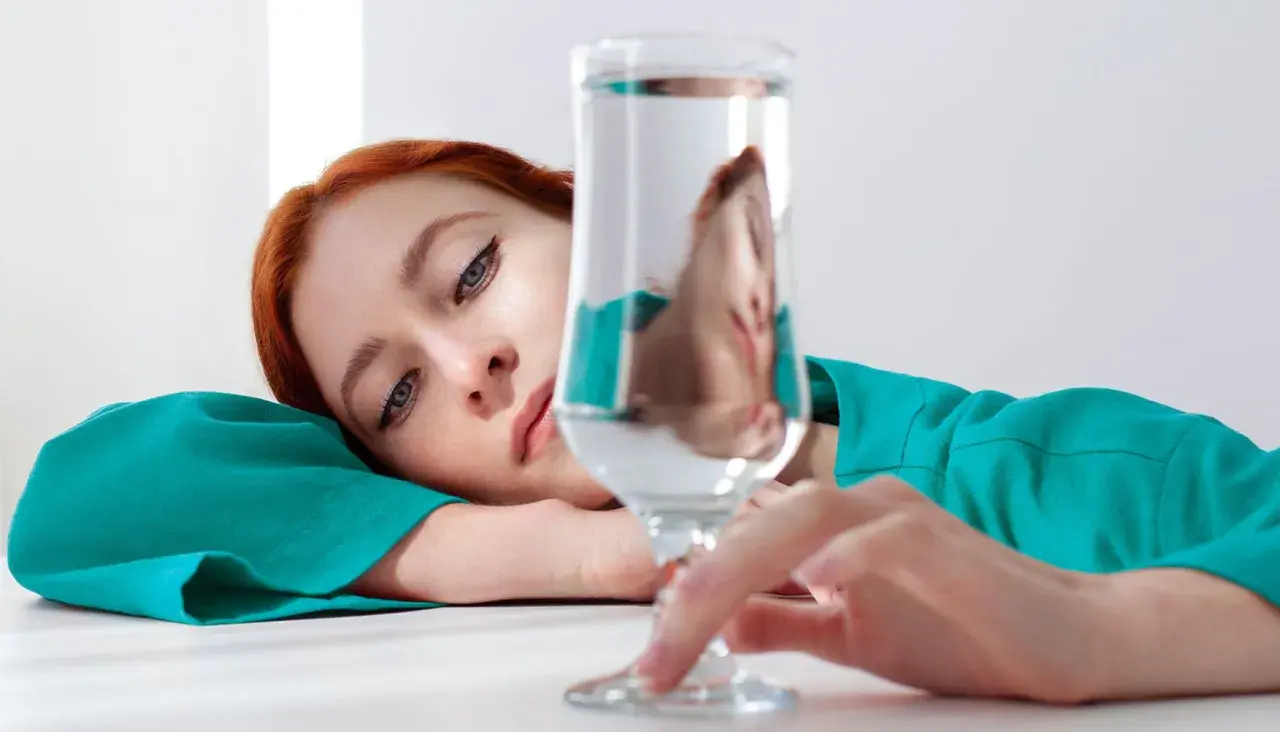 Mujer pelirroja recostada, con su rostro reflejado en un vaso de agua, quizás pensando en los beneficios de un alcalinizador de agua para su bienestar.