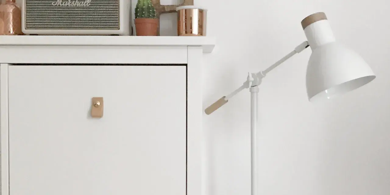 Un mueble blanco con un tirador de cuero y un altavoz Marshall. Una lámpara blanca con detalles de madera ilumina la escena.