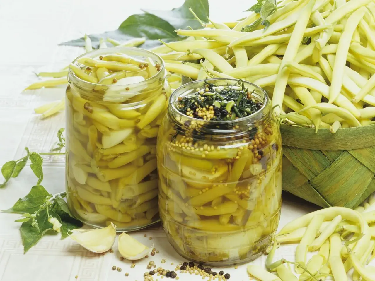 Zwei Gläser mit eingelegten Buschbohnen, daneben frische Bohnen und Knoblauch.