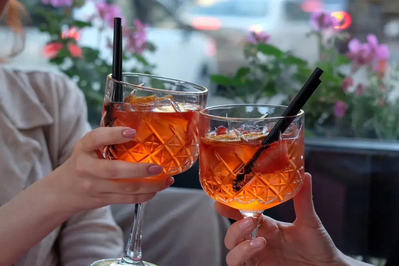 Dwie osoby wznoszą toast Aperol Spritz, ozdobionymi owocami i słomkami, na tle kwitnących kwiatów.