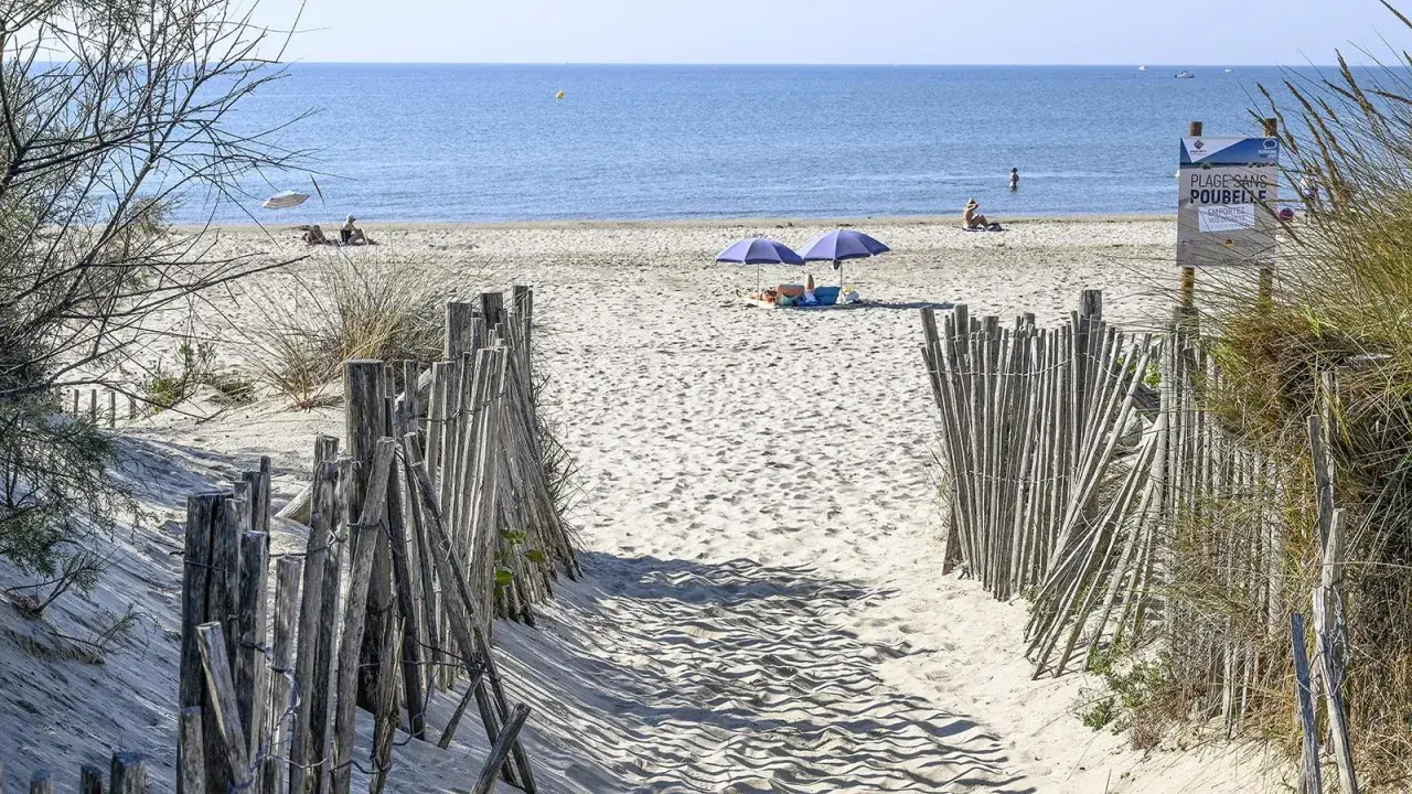 Ein sandiger Weg führt zum Montpellier Strand. Zwei Sonnenschirme spenden Schatten, während Menschen die Sonne genießen.