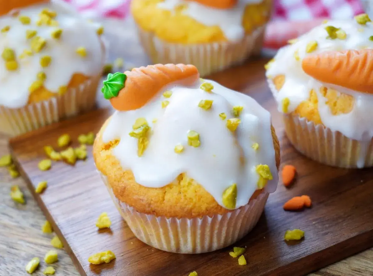 Muffins mit weißer Glasur, gehackten Pistazien und Marzipan-Karotten auf Holzbrett.
