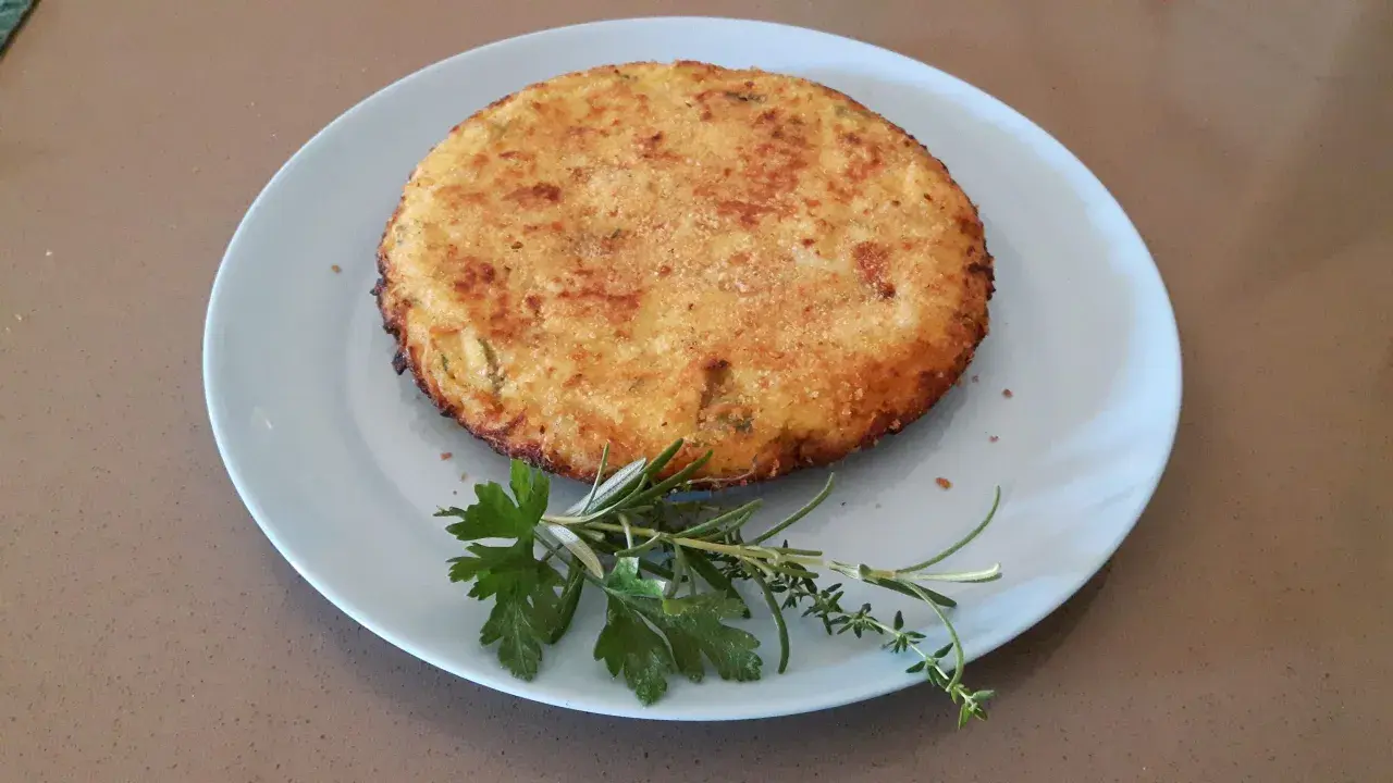 Frittata di cous cous dorata e croccante, guarnita con erbe aromatiche fresche su un piatto azzurro.