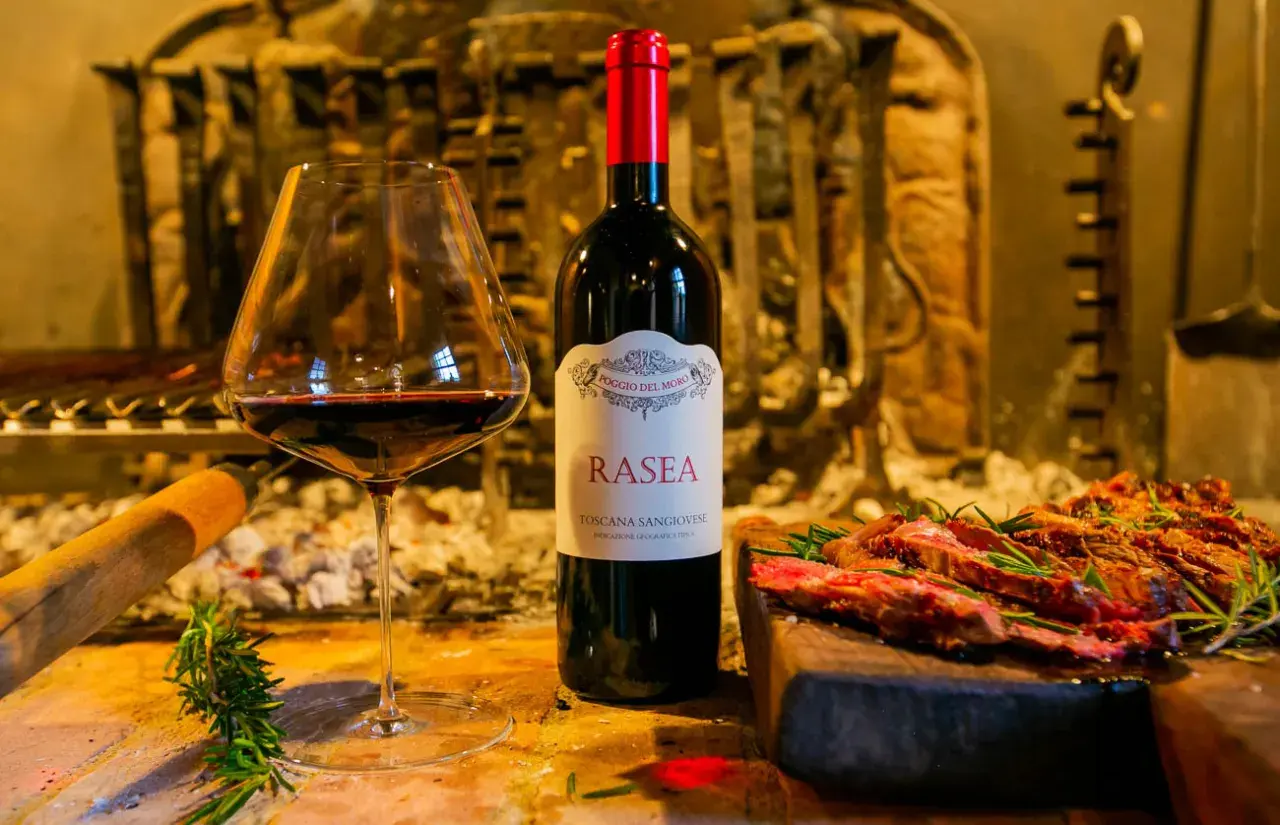 Calice di vino rosso accanto a una bottiglia di Rasea, un vino toscano, con carne grigliata e rosmarino. Un'ode ai super tuscan wines.