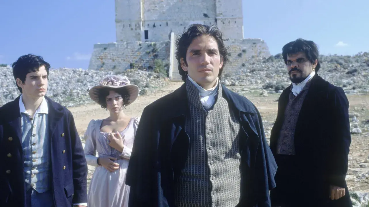 Młody mężczyzna w stroju z epoki, jak z filmu hrabia Monte Cristo 2024, stoi na tle kamiennej budowli. Obok niego kobieta w kapeluszu i inny mężczyzna.