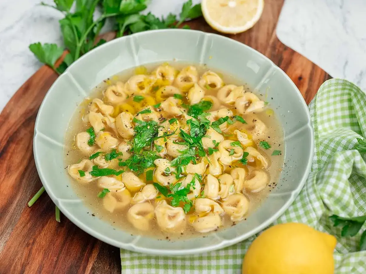 En skål med tortellini in brodo, garnerad med färsk persilja och en citronskiva.