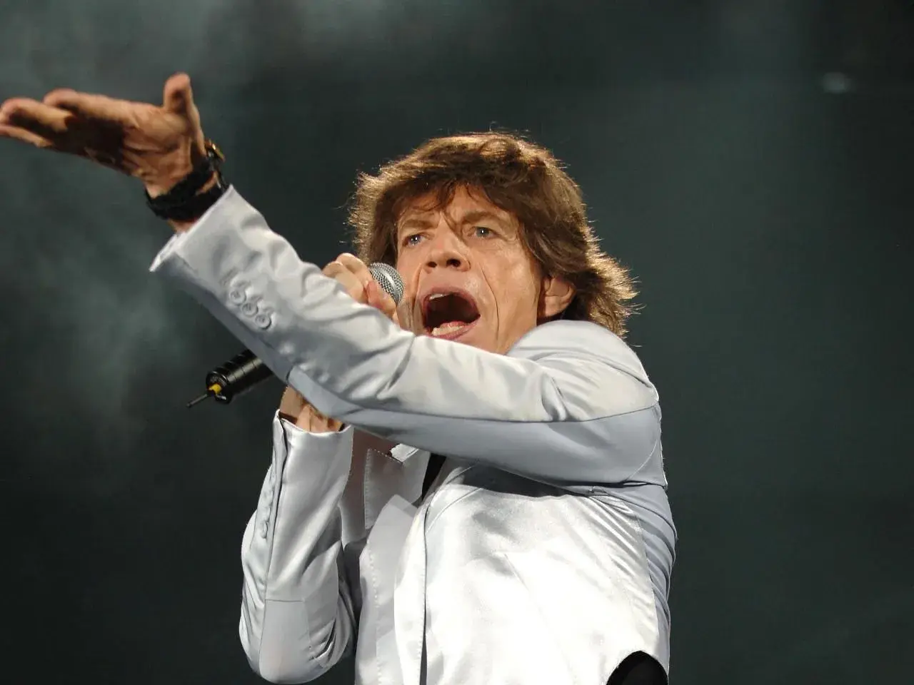 Mick Jagger śpiewa, wykonując swoje legendarne utwory. Wokalista w srebrnej marynarce z mikrofonem w dłoni.
