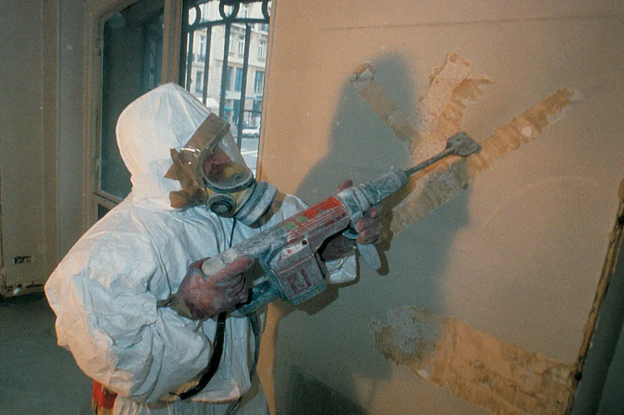 Travailleur en combinaison intégrale et masque à gaz, utilisant un marteau-piqueur pour retirer un revêtement mural. Que faire face à l'amiante au travail.