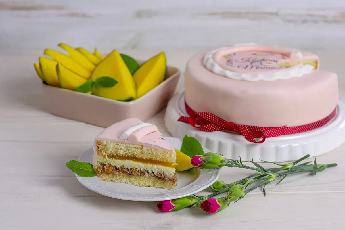 Różowy tort z napisem "Kocham Mamę", udekorowany bielą tytanową spożywczą, obok kawałek ciasta i mango.