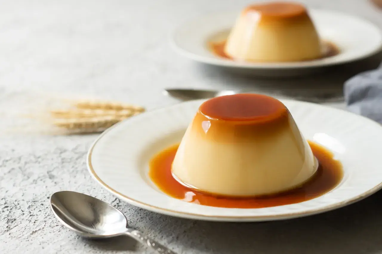 Zwei cremige flan kuchen mit flüssiger Karamellsoße auf weißen Tellern, daneben Löffel und ein paar Ähren.