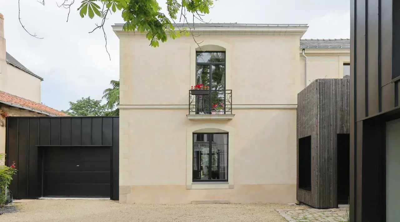 Maison rénovée par un architecte DPLG, mêlant pierre claire, bois sombre et garage noir moderne. Balcon fleuri sur la façade.