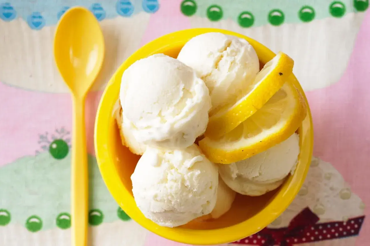 Erfrischendes Buttermilch Zitronen Eis in einer gelben Schale, garniert mit Zitronenscheiben. Eine gelbe Löffel liegt daneben.