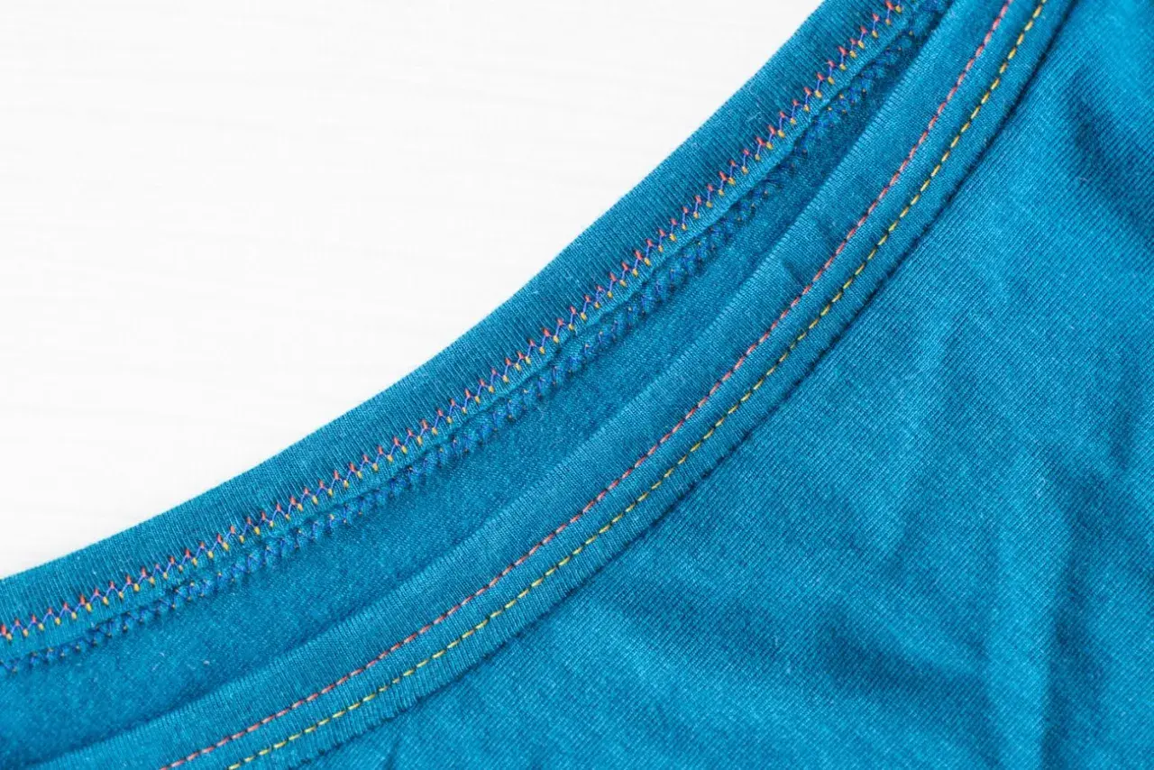 Close-up of teal fabric showing a decorative double needle topstitch with colorful threads, adding a vibrant detail to the garment's edge.