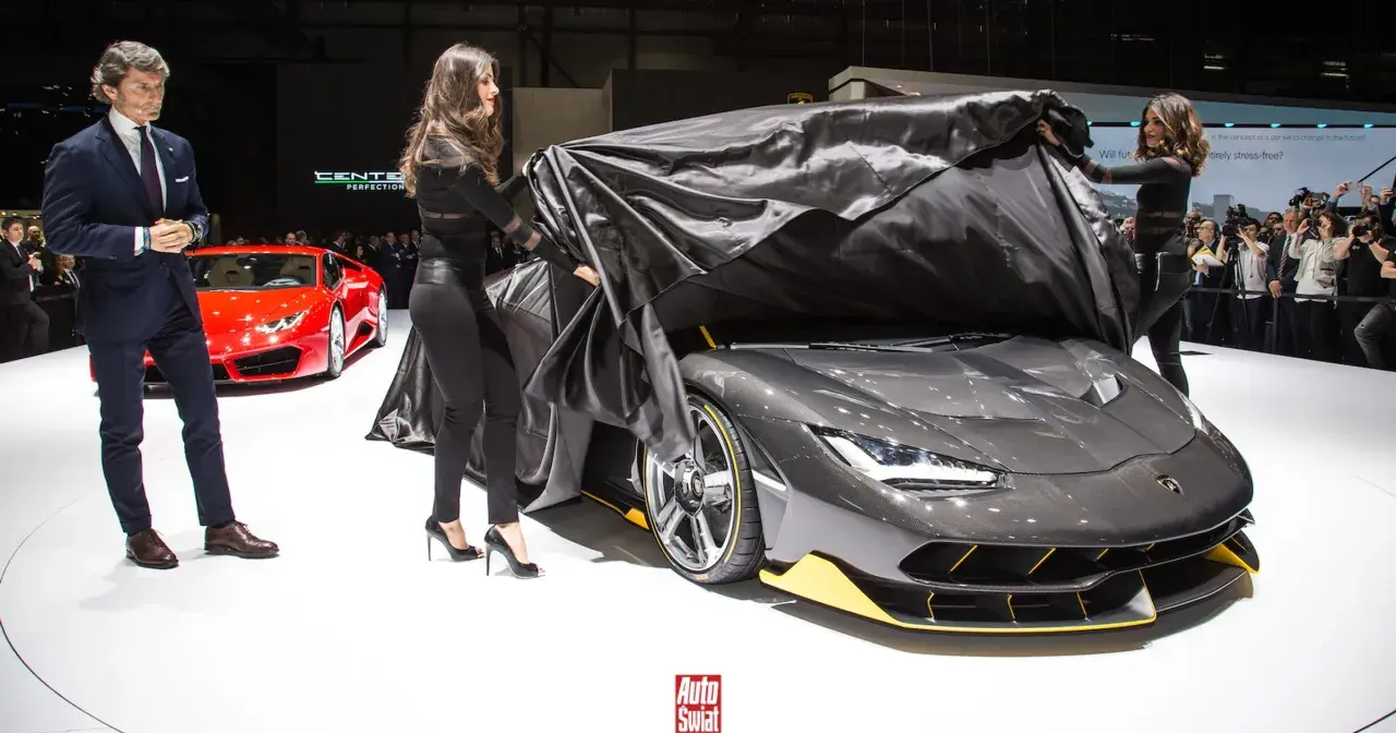 Elegancki mężczyzna i dwie kobiety odsłaniają nowy, szary Lamborghini Centenario. Obok stoi czerwony sportowy samochód.
