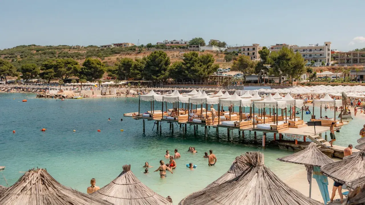 Słoneczny dzień na plaży Ksamil. Ludzie kąpią się w turkusowej wodzie, odpoczywają pod białymi parasolami na pomostach i na piaszczystym brzegu.