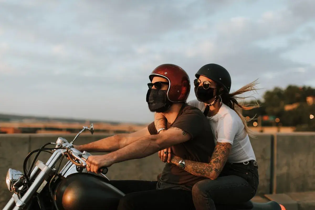 Pareja con mascarillas, montando a horcajadas moto al atardecer. El viento agita el cabello de ella.