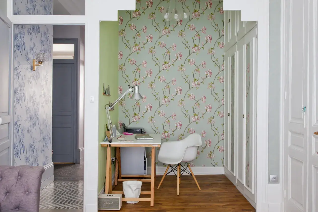 Un pan de mur de salon est habillé d'un papier peint floral aux fleurs roses sur fond vert pâle. Un bureau et une chaise blanche complètent l'espace.