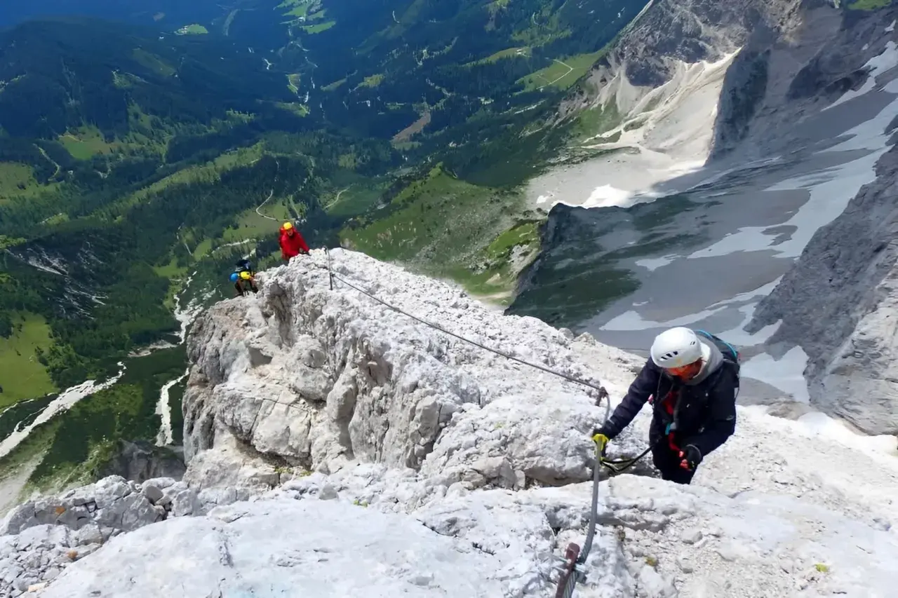 Escaladores en una vía ferrata, asegurados a un cable de acero que recorre la cresta rocosa. El paisaje montañoso es espectacular.