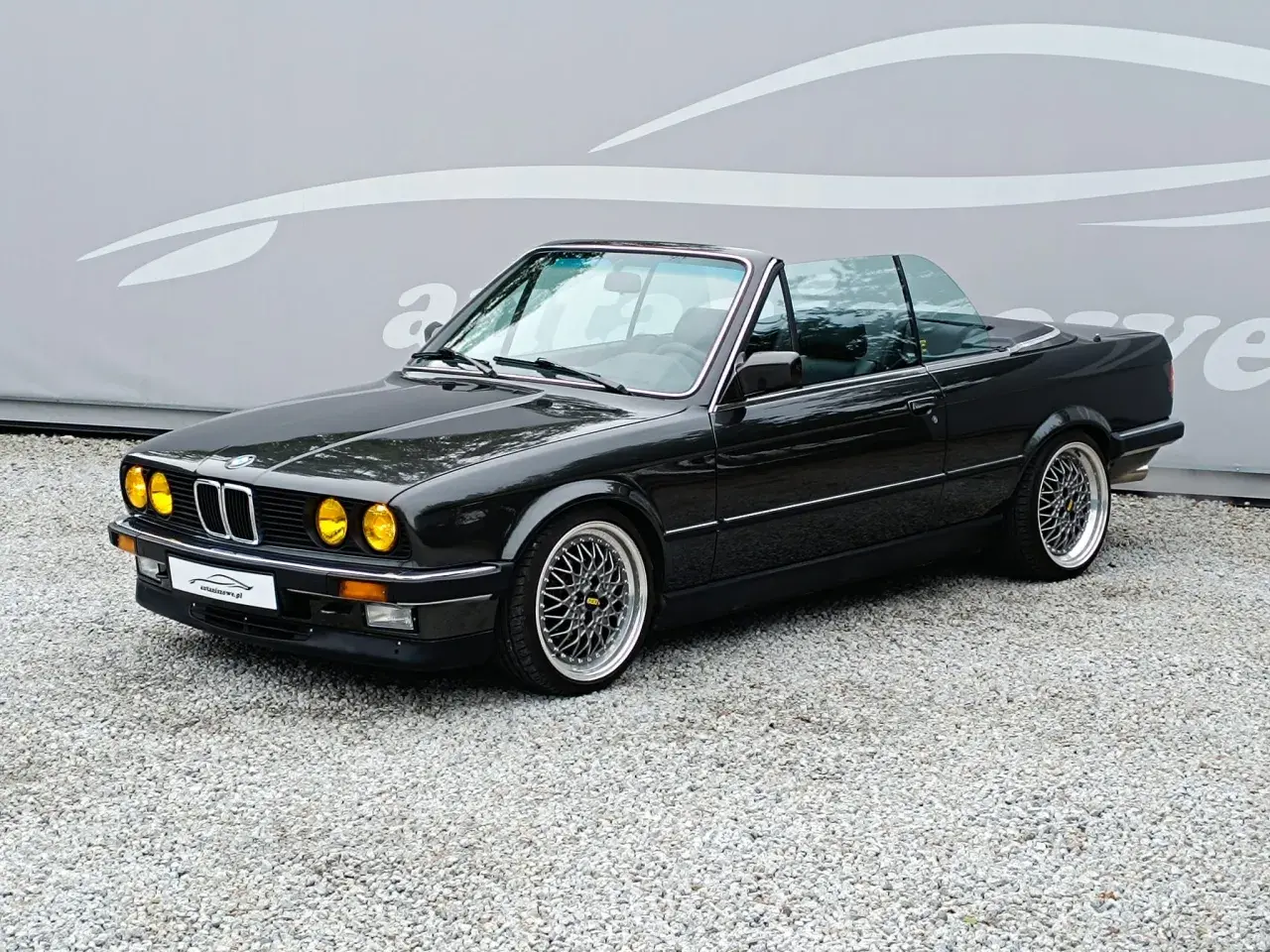 Czarny kabriolet BMW E30 z żółtymi światłami i felgami BBS. Klasyk na drodze.