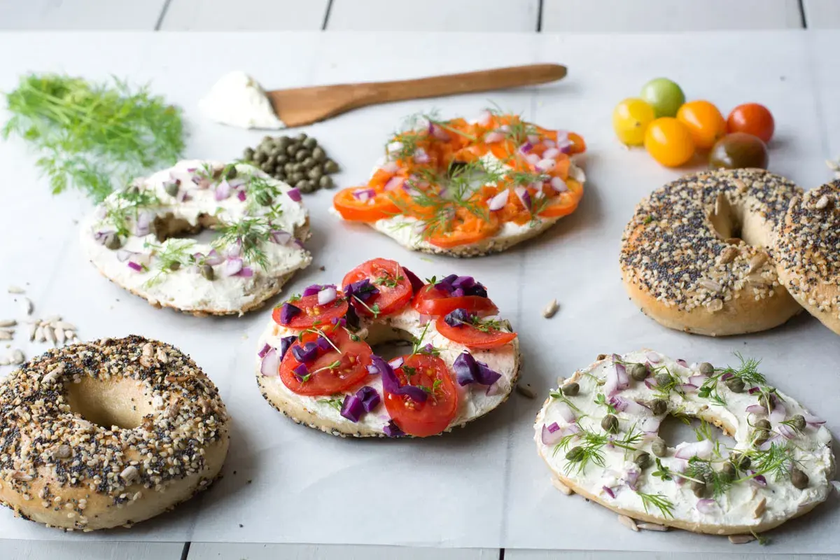 Flera bröd med hål i mitten, toppade med färskost, lax, tomater, rödlök och dill. Ett härligt urval av bagels!
