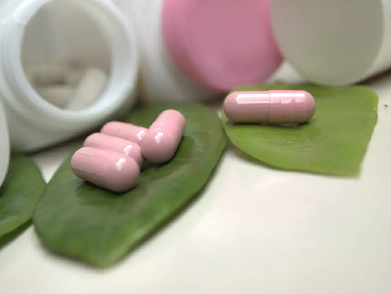 Różowe kapsułki, być może zawierające ketoprofen, leżą na zielonych liściach obok otwartych pojemników z lekami.