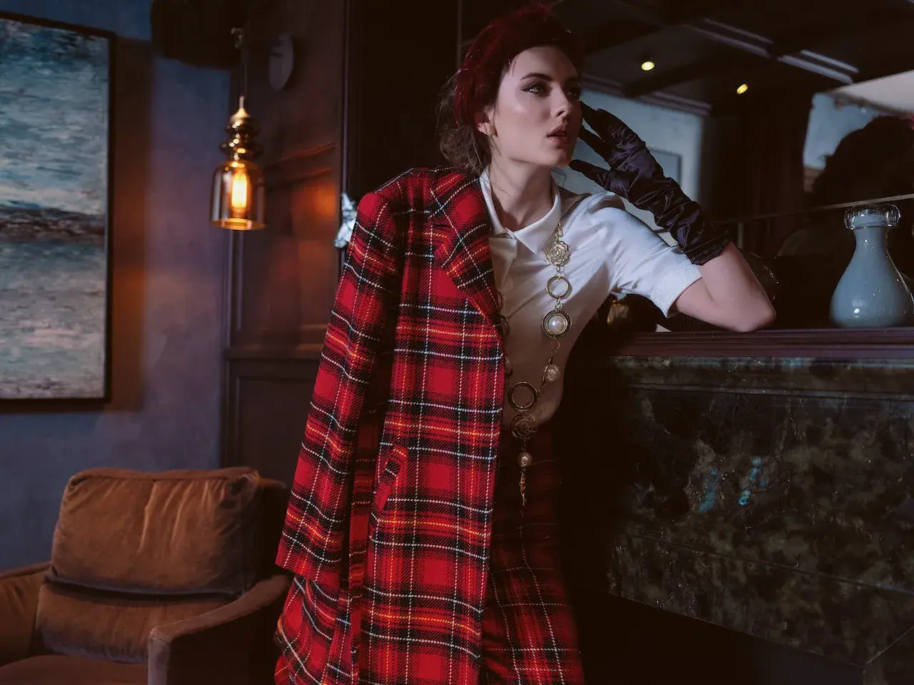 Une femme pose, vêtue d'un blazer au **motif tartan** rouge et noir, avec un collier perlé.