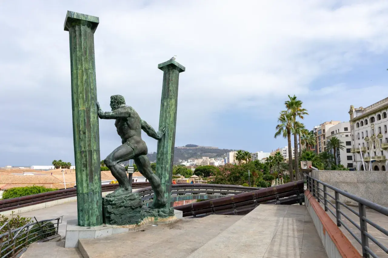 Statua di Ercole che sorregge le colonne d'Ercole oggi, con palme e edifici sullo sfondo.