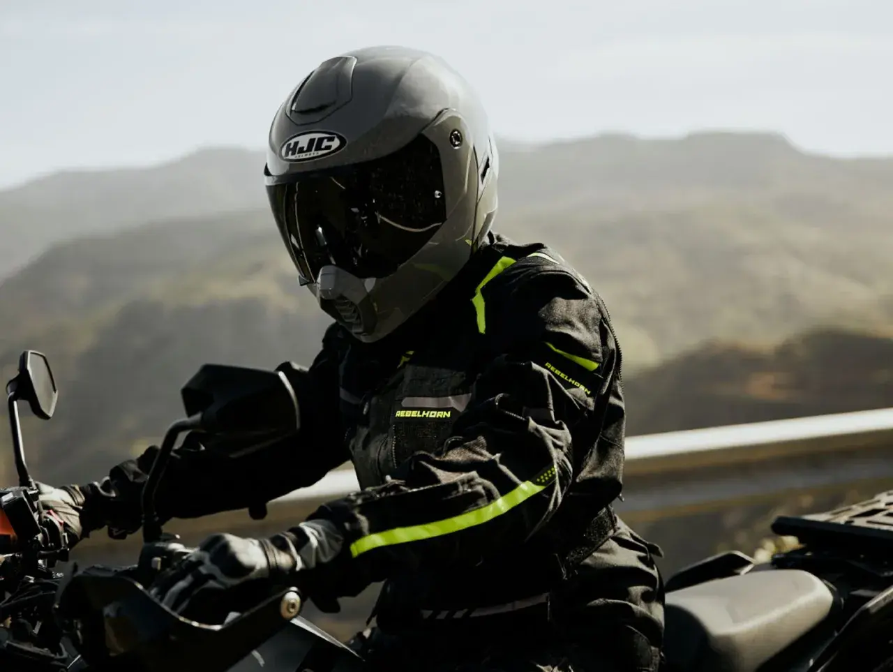 Motocyklista w szarym kasku HJC i czarnej kurtce z neonowymi detalami jedzie motocyklem po górskiej drodze.