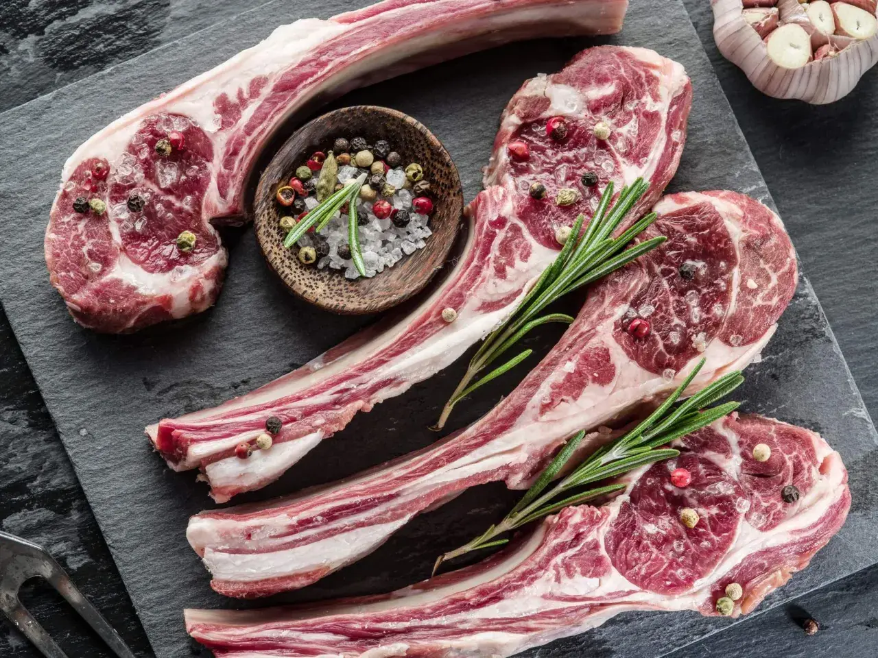 Côtelettes d'agneau crues, assaisonnées de sel et de poivre, prêtes pour une recette d'agneau pascal. Romarin frais et ail complètent la scène.