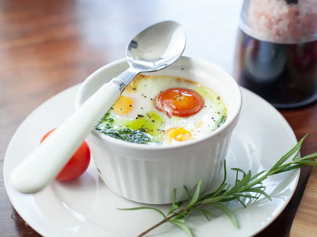 Un œuf ramequin cuit au four, garni d'une tomate et d'herbes, prêt à être dégusté avec une cuillère.