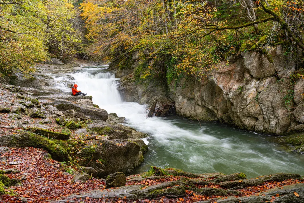 Persona sentada en rocas junto al Saut deth Pish, rodeada de árboles otoñales y agua turbulenta.