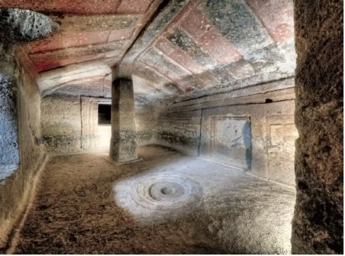 Interno di una tomba etrusca con soffitto dipinto e un pilastro centrale. Un cerchio inciso sul pavimento sembra un antico incantesimo putifigari.