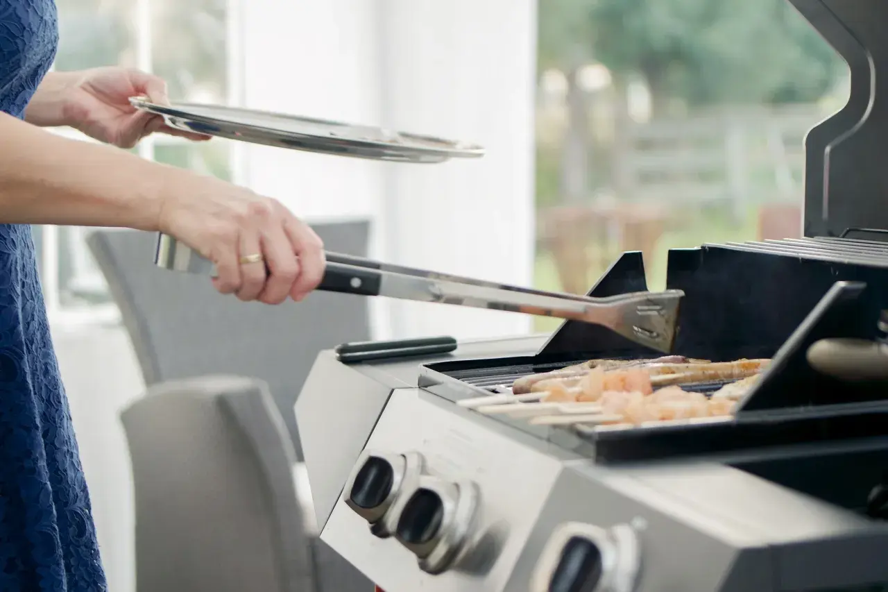 Person i blå klänning använder tång för att vända kycklingspett på en fyrkantig kolgrill.