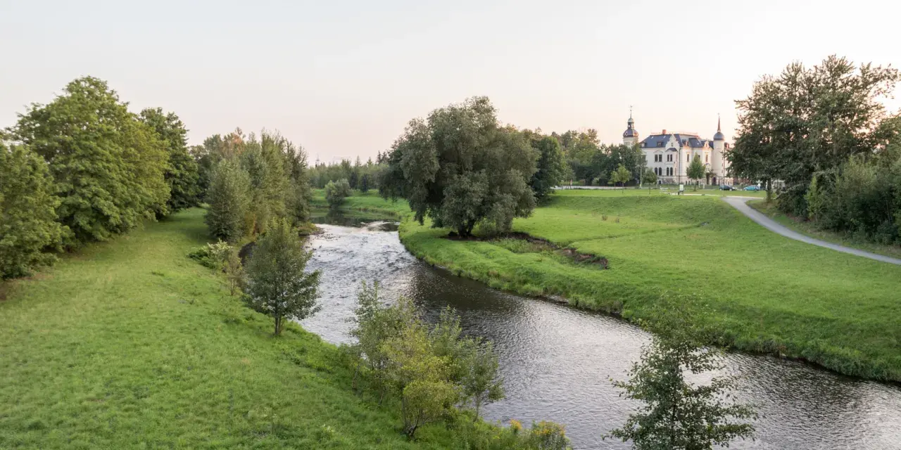 Idyllischer Flusslauf mit grünen Ufern und einem Schloss im Hintergrund. Ein perfekter Ort für Radwege Geheimtipp-Touren.